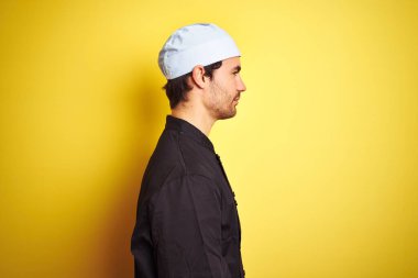 Üniformalı ve şapkalı genç, yakışıklı aşçı yalıtılmış sarı arka planda, rahat profil pozu veren, kendine güveni tam bir gülümseme takınıyor..