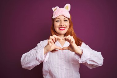 Genç, güzel, kızıl saçlı, pijama giyip maske takan, mor, soyutlanmış bir geçmişi olan bir kadın. Aşkla gülümseyen, elleriyle kalp sembolü gösteren bir kadın. Romantik konsept.