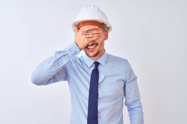 Müteahhit kaskı takan genç bir iş adamı izole edilmiş arka planda gülümsüyor ve yüzünde şaşkınlıktan gözleri kapayarak gülüyor. Kör kavram.