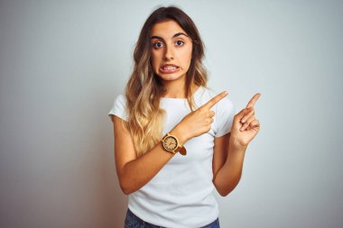 İzole edilmiş arka plan üzerinde gündelik beyaz tişört giyen genç ve güzel bir kadın iki eliyle endişeli ve endişeli bir ifadeye işaret ediyor.