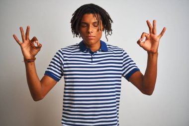 Rasta desenli mavi polo giyen Afro bir adam izole edilmiş beyaz arka plan üzerinde duruyor rahatlamış ve gözleri kapalı meditasyon hareketi yaparken parmaklarıyla gülümsüyor. Yoga konsepti.