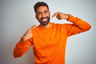 İzole edilmiş beyaz arka planda turuncu süveter giyen genç Hintli adam gülümseyen ve parmaklarıyla ve ağzıyla işaret eden. Diş sağlığı konsepti.
