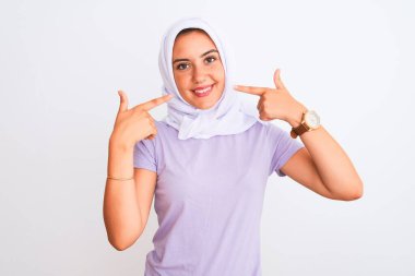 İzole edilmiş beyaz arka planda tesettürlü, tesettürlü, neşeli gülümseyen ve parmaklarıyla ve ağzıyla işaret eden genç, güzel bir Arap kızı. Diş sağlığı konsepti.