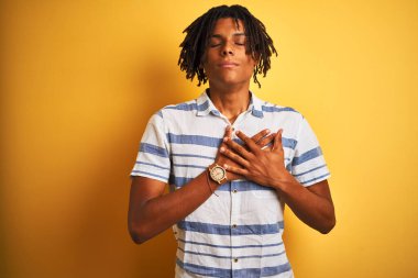 Sarı arka plan üzerinde çizgili tişört giyen, elleri göğsünde, gözleri kapalı ve yüzünde minnet dolu bir jest olan Afro-Amerikan bir adam. Sağlık kavramı.