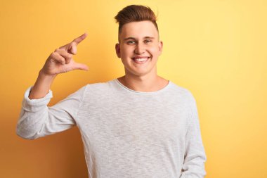 İzole edilmiş sarı arka plan üzerinde duran beyaz tişörtlü genç yakışıklı adam gülümsüyor ve kendine güvenen el hareketleriyle küçük beden işareti yapıyor ve kameraya bakıyor. Ölçü kavramı.