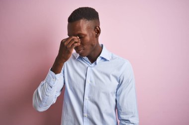İzole edilmiş pembe arka plan üzerinde duran mavi şık gömlekli Afro-Amerikan bir adam yorgun burnu ve gözleri ovuşturarak yorgunluk ve baş ağrısı hissediyor. Stres ve hüsran kavramı.