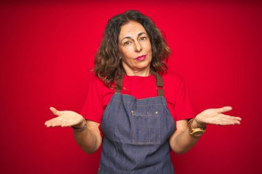 Kırmızı izole arka planda önlük giyen orta yaşlı bir kadın hiçbir şeyden habersiz ve kolları açık, hiçbir fikri yok..