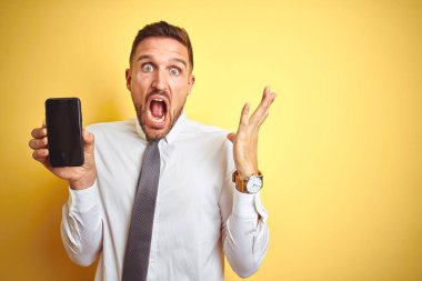 Genç yakışıklı iş adamı sarı izole edilmiş arka planda akıllı telefon ekranı gösteriyor. Çok mutlu ve heyecanlı, kazanan yüz ifadesi zaferi kutluyor. Büyük bir gülümsemeyle ve el kaldırarak.