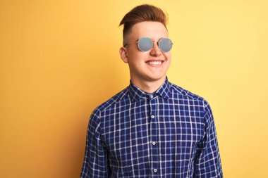 Sarı arka planda güneş gözlüğü ve günlük gömlek takan yakışıklı genç adam yüzünde gülümseme, doğal bir ifade ile yan yana bakıyor. Kendinden emin gülen.
