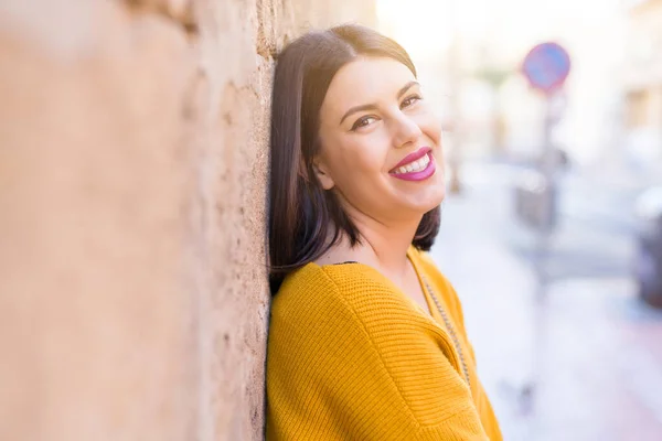 Beautiful young woman smiling confident and cheerful leaning on bricks wall, walking on the street of the city on a sunny day