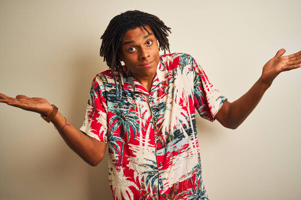 Afro man with dreadlocks on vacation wearing summer shirt over isolated white background clueless and confused expression with arms and hands raised. Doubt concept.