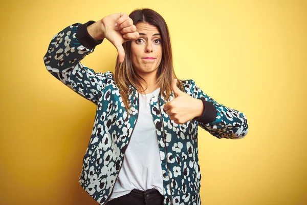 Young beautiful woman wearing casual jacket over yellow isolated background Doing thumbs up and down, disagreement and agreement expression. Crazy conflict