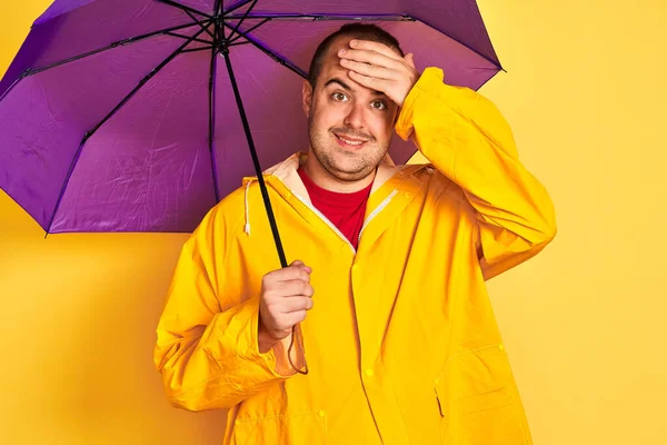 Young man wearing raincoat holding purple umbrella standing over isolated yellow background stressed with hand on head, shocked with shame and surprise face, angry and frustrated. Fear and upset for mistake.