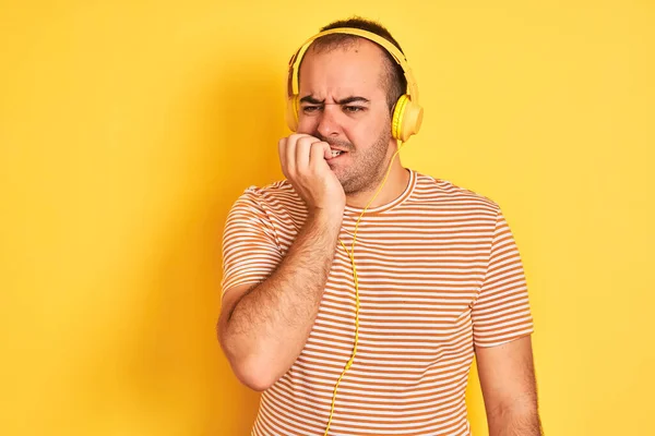 Young man listening to music using headphones standing over isolated yellow background looking stressed and nervous with hands on mouth biting nails. Anxiety problem.