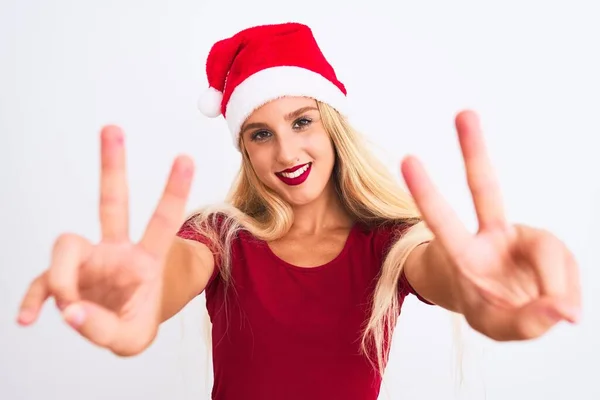 孤立した白い背景の上にクリスマスのサンタの帽子を身に着けている若い美しい女性は勝利の印をしている指を示すカメラを見て笑っている 第二番 — ストック写真