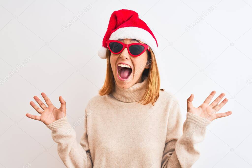 Beautiful redhead woman wearing christmas hat and red sunglasses very happy and excited, winner expression celebrating victory screaming with big smile and raised hands