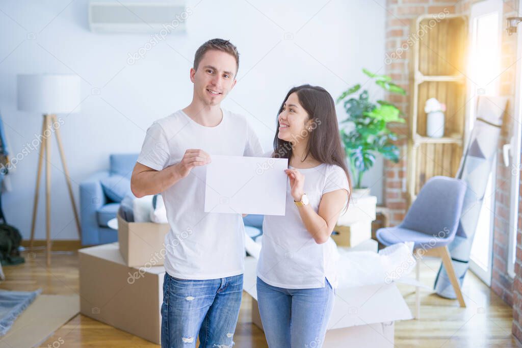 Young beautiful couple holding banner at new home around cardboard boxes