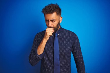 Zarif gömlek ve kravat giyen genç Hintli işadamı izole edilmiş mavi arka planda dikiliyor ve soğuk algınlığı veya bronşitin belirtisi olarak öksürüyor. Sağlık hizmetleri kavramı.