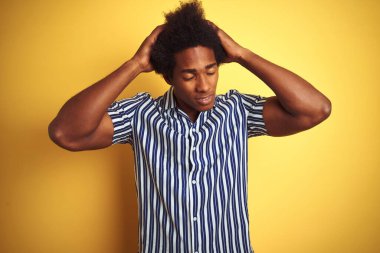 Afro saçlı, çizgili tişörtlü Amerikalı bir adam izole edilmiş sarı arka plan üzerinde duruyor. Ağrı ve migren yüzünden baş ağrısı ve stresten muzdarip. Ellerini başının üstüne koy..