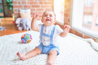 Güzel bebek battaniyenin üzerinde oturuyor. Anaokulunda elleri havada gülümsüyor.
