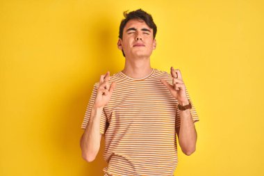 Teenager boy wearing yellow t-shirt over isolated background gesturing finger crossed smiling with hope and eyes closed. Luck and superstitious concept. clipart
