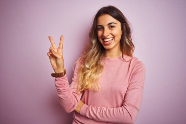 Pembe izole edilmiş arka planda süveter giyen genç ve güzel bir kadın kameraya gülümseyen bir gülümsemeyle zafer işareti yapıyor. İki numara..