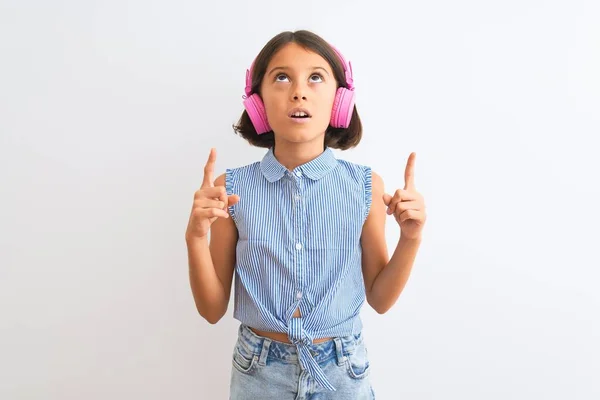 美しい子供の女の子は 孤立した白い背景の上にヘッドフォンを使用して音楽を聞いて驚いて指と上げ腕で見上げと指摘 — ストック写真