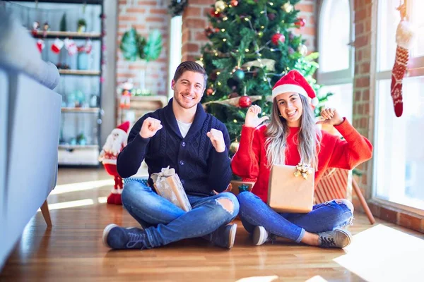 这对年轻夫妇头戴圣爪帽 坐在圣诞树旁的地板上 满脸笑容 满脸自信 用手指指着自己 自豪而快乐 — 图库照片