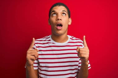 İzole edilmiş kırmızı arka plan üzerinde çizgili tişört giyen genç yakışıklı Arap adam şaşırmış ve şaşırmış bir şekilde yukarı bakıyor ve parmaklarıyla işaret ediyor ve kollarını kaldırıyordu..