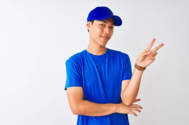 Mavi tişört ve kep giyen Çinli teslimatçı izole edilmiş beyaz arka planda dikiliyor ve kameraya gülümsüyor zafer işareti yapıyor. İki numara..