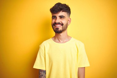 Dövmeli, tişörtlü genç bir adam izole edilmiş sarı arka plan üzerinde duruyor. Yüzünde gülümseme, doğal bir ifade ile yan yana bakıyor. Kendinden emin gülen.