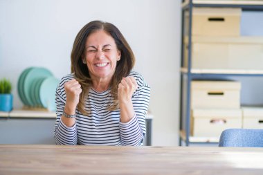 Orta yaş kıdemli kadın evde masada oturan silah ile başarı için heyecanlı kaldırdı ve gözleri zafer gülümseyen kutluyor kapalı. Kazanan kavramı.