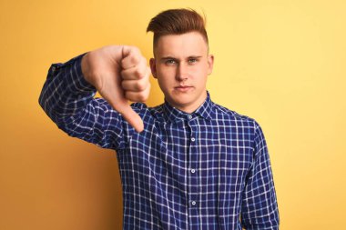 Genç yakışıklı adam izole sarı arka plan üzerinde duran rahat gömlek giyen mutsuz ve kızgın gösteren reddi gösteren ve aşağı jest başparmak ile negatif. Kötü ifade.