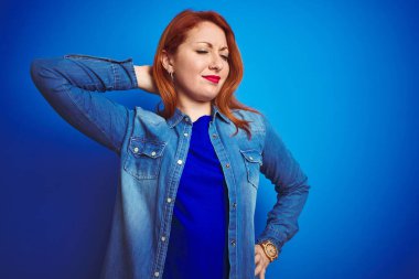Genç, kızıl saçlı, kot gömlek giyen, mavi izole edilmiş arka planda duran, boynu ağrıyan, eliyle boynuna dokunan, kasları ağrıyan bir kadın.