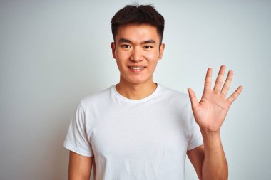 Asyalı genç bir Çinli tişört giyiyor. İzole edilmiş beyaz arka plan üzerinde duruyor. Beş numarayı işaret ediyor ve gülümsüyor..