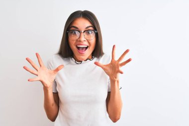 Genç ve güzel bir kadın günlük tişört ve gözlük takıp izole edilmiş beyaz arka planda çılgınlığı kutluyor ve başarıya hayret ediyor. Kolları havada ve gözleri açık, heyecanlı bir şekilde çığlık atıyor. Kazanan konsept