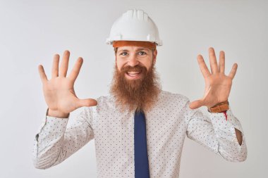 İzole edilmiş beyaz arka plan üzerinde güvenlik kaskı takan genç kızıl saçlı mimar kendini beğenmiş ve mutlu bir şekilde gülümserken on numarayı işaret ediyor..