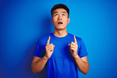 İzole edilmiş mavi arka planın üzerinde duran Asyalı genç adam şaşırdı ve şaşırdı. Parmaklarıyla işaret etti ve kollarını kaldırdı..