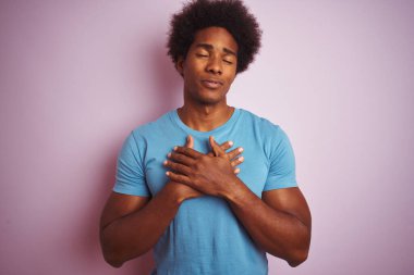 Afro saçlı, mavi tişörtlü, izole edilmiş pembe arka planda duran, elleri göğsünde, gözleri kapalı ve yüzünde minnet dolu bir jest olan Afro-Amerikan bir adam. Sağlık kavramı.