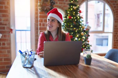 Noel 'de dizüstü bilgisayarla çalışan güzel bir kadın Noel Baba şapkası takıyor ve yüzünde gülümseme, doğal bir ifade ile yan yana bakıyor. Kendinden emin gülen.