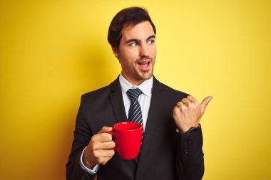 Genç yakışıklı işadamı izole edilmiş sarı arka planda kırmızı kahve içerken başparmağıyla gülümsüyor.