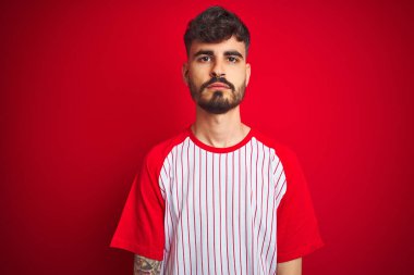 Yüzünde ciddi bir ifade olan, izole edilmiş kırmızı arka plan üzerinde duran çizgili tişörtlü genç bir adam. Basit ve doğal bir şekilde kameraya bakıyorsun..