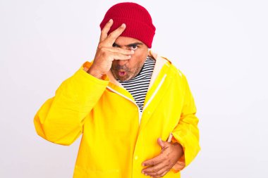 Yağmurluk giyen ve yünlü şapka takan orta yaşlı bir adam izole edilmiş beyaz arka planda duruyor. Şok olmuş bir şekilde yüzünü ve gözlerini kapıyor, utanmış bir ifadeyle parmaklarından bakıyor..