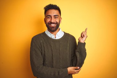 Yeşil kazak ve gömlek giyen genç Hintli adam izole edilmiş sarı arka planda duruyor. Yüzünde kocaman bir gülümseme var. Eli ve parmağı kameraya bakıyor..