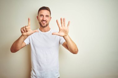 Gösterilen ve parmakları ile işaret izole arka plan üzerinde rahat beyaz t-shirt giyen genç yakışıklı adam kendinden emin ve mutlu gülümseyerek süre yedi numara.