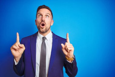 Genç ve yakışıklı iş adamı mavi arka planda şaşkın ve şaşırmış görünüyor ve parmaklarıyla işaret ediyor ve kollarını kaldırıyor..