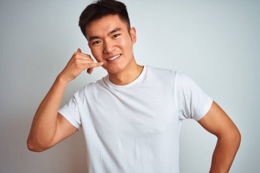 Asyalı genç bir Çinli tişört giyip izole edilmiş beyaz arka planda duruyor, gülümsüyor telefonla el ve parmaklarıyla telefon hareketi yapıyor, telefonda konuşuyor gibi. İletişim kavramları.