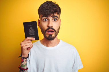 Kanada pasaportu takan genç bir adam izole edilmiş sarı arka plan. Şaşırmış bir yüzle korkmuş ve korku ifadesinden heyecanlanmış.