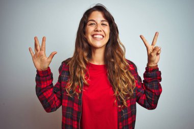 Kırmızı tişört ve ceket giyen genç güzel bir kadın beyaz izole edilmiş arka plan üzerinde duruyor ve yedinci parmaklarıyla işaret ederken kendinden emin ve mutlu bir şekilde gülümsüyor..