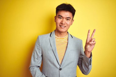 İzole edilmiş sarı arka planda duran ceketli genç Asyalı iş adamı kameraya bakıp gülümsüyor ve zafer işareti yapıyor. İki numara..